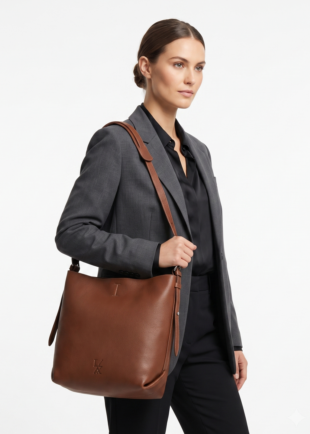 A woman in a dark gray blazer and black outfit models a brown leather shoulder bag with a matching strap against a clean white background.