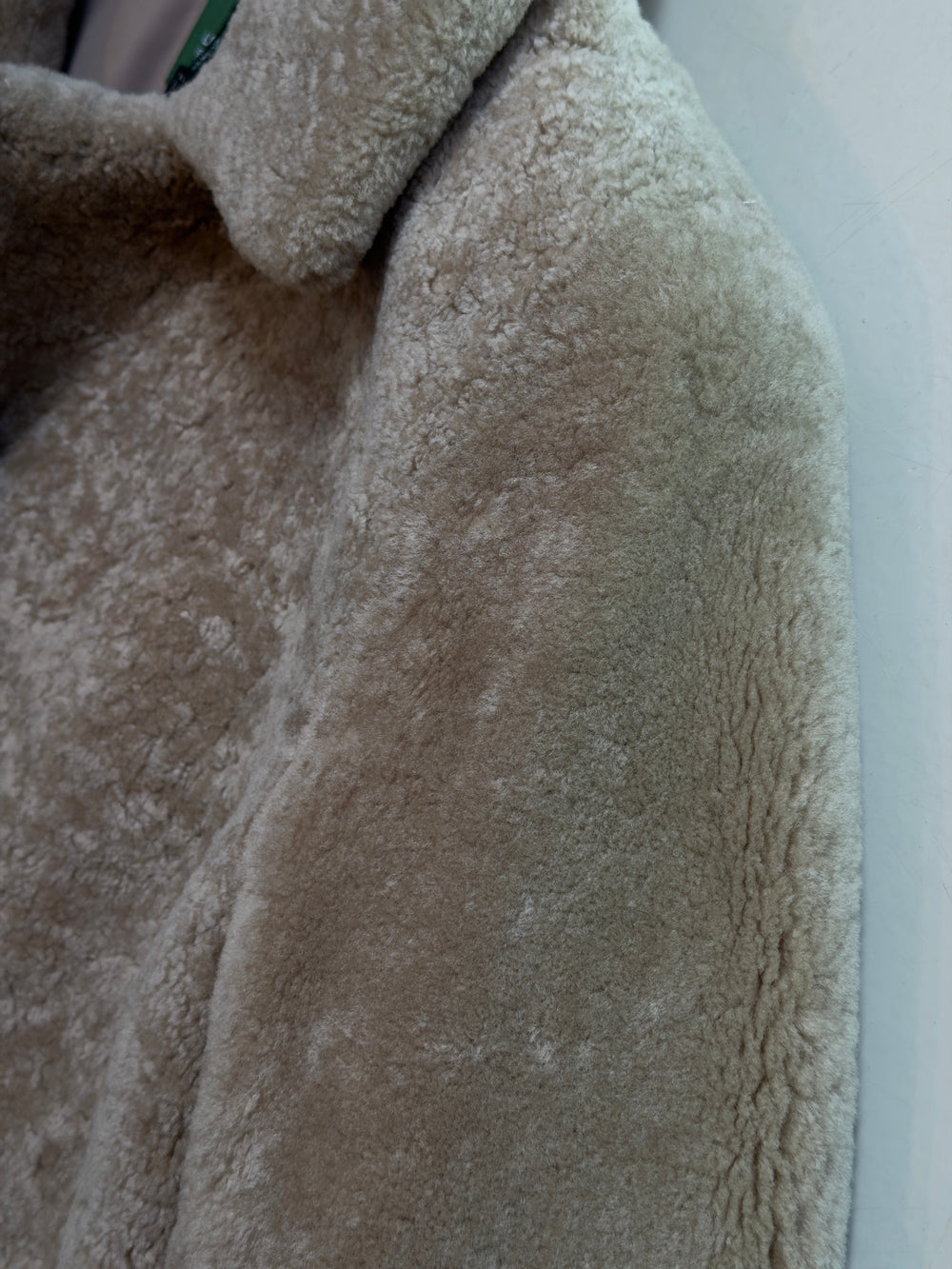 Close-up of a plush, textured faux-shearling or sherpa jacket in a warm taupe-beige color, showing the soft pile fabric and collar detail.