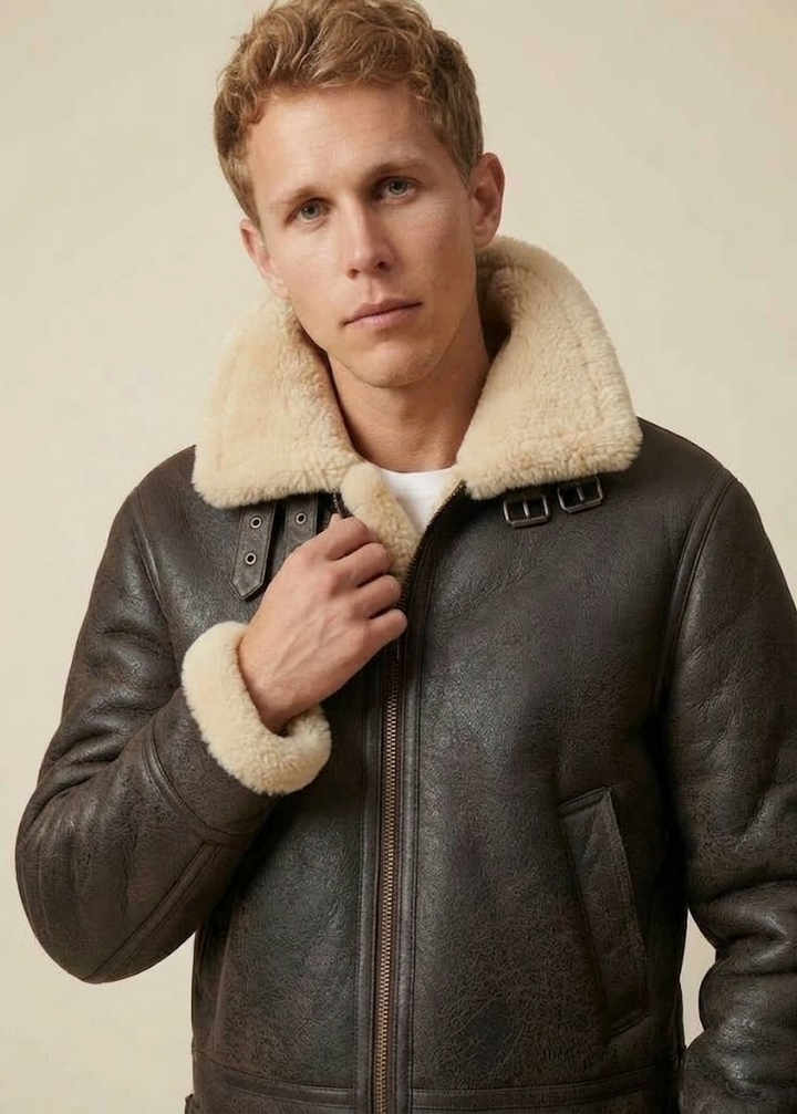 A man wearing a dark brown leather aviator-style jacket with a thick cream shearling collar and cuffs, photographed against a plain beige studio background.