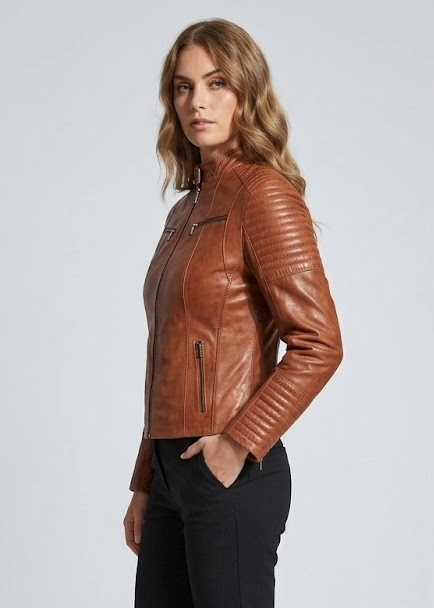 A woman modeling a brown leather moto jacket with quilted sleeves, paired with black trousers and black ankle boots, standing against a light gray studio background.