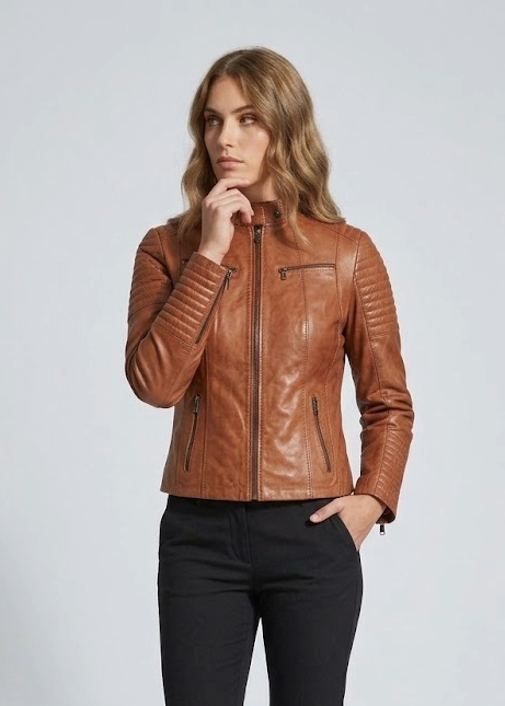 A woman modeling a cropped brown leather moto jacket with black pants and black ankle boots, standing against a light gray studio background.
